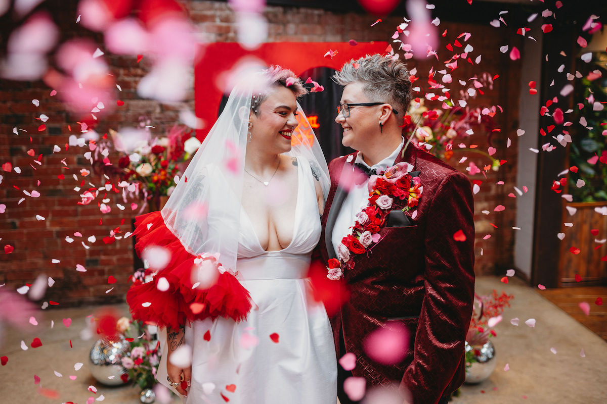 Two brides stand together as red and pink confetti rain down on them