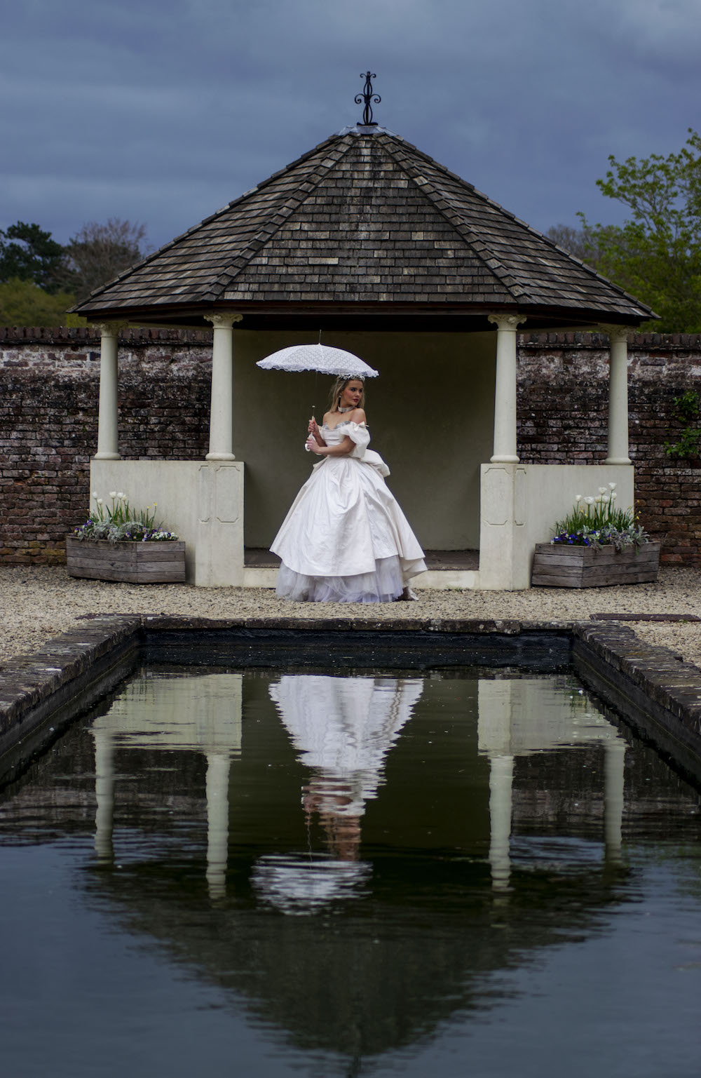 bride holding a parasol in front of a pergola with her reflection in the lake