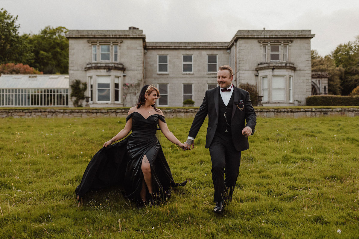 bride wearing black wedding dress holds her grooms hand as they walk across a field in front of their Cornwall wedding venue