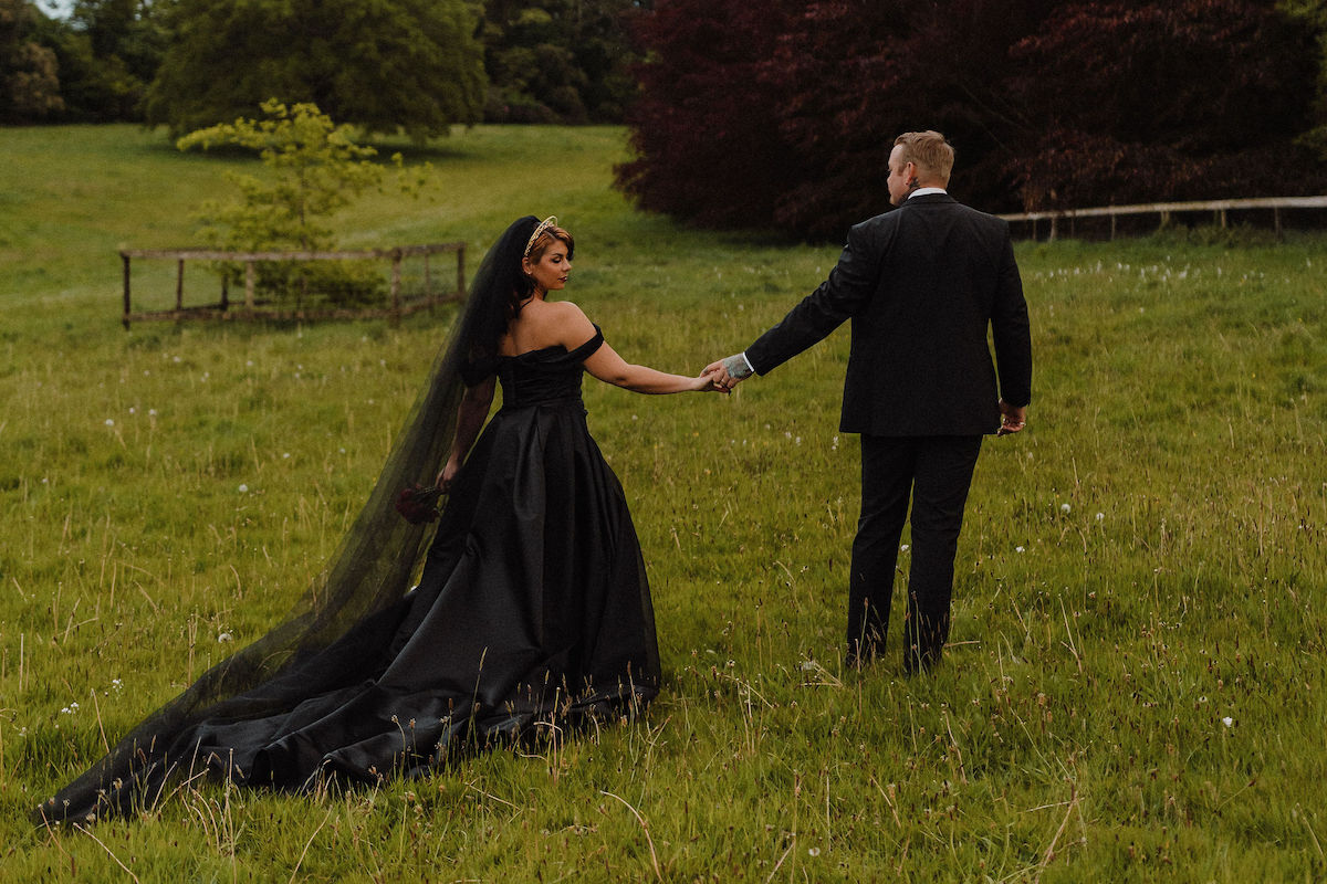 bride wearing black wedding dress holds her grooms hand as they walk across a field in Cornwall