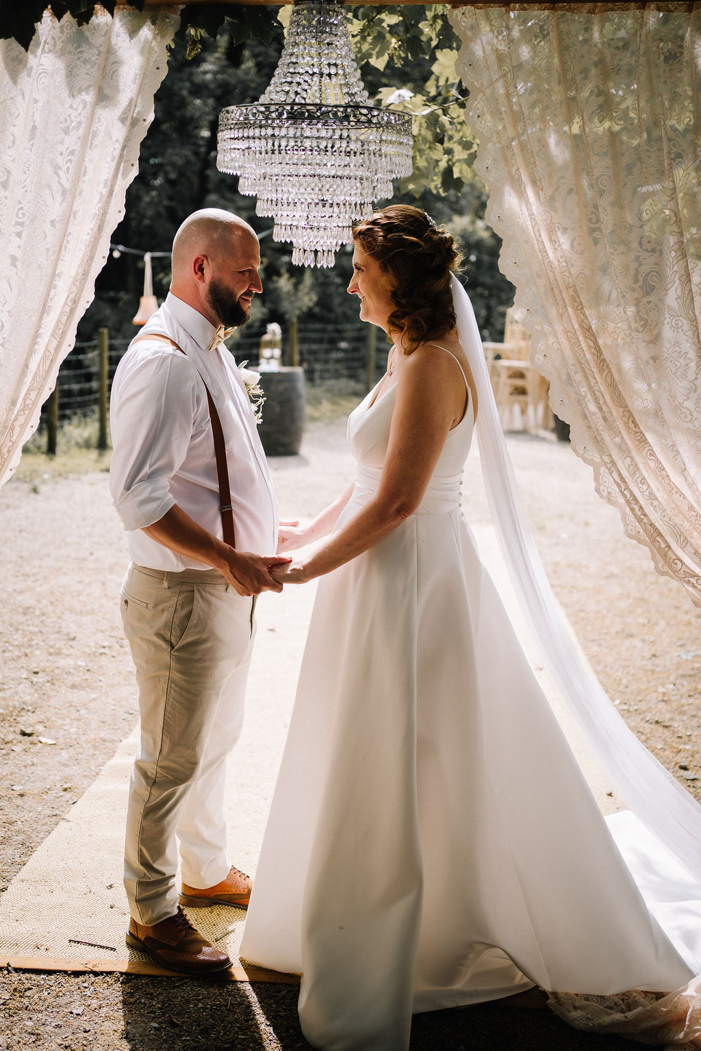 bride and groom stand under a vintage chandelier surrounded by vintage lace curtains making the ceremony area