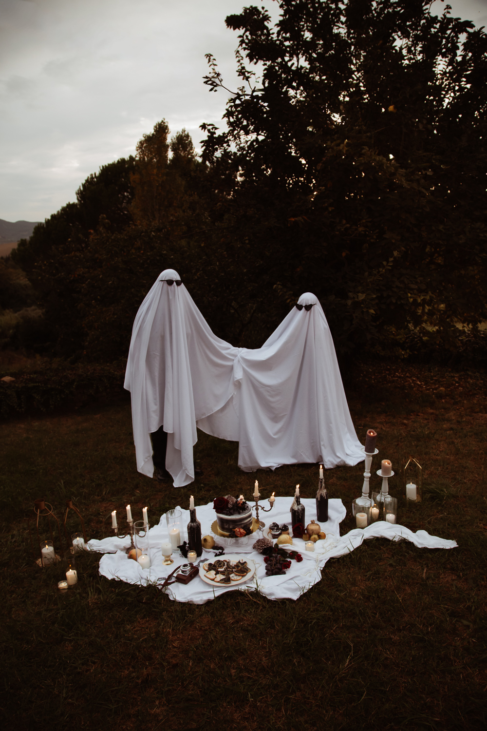 couple dressed as ghosts wearing sunglasses holding hands as the sun sets for halloween portrait
