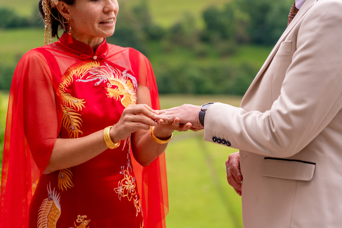 close up of bride wearing Chinese wedding dress placing the wedding ring on her grooms finger